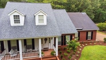 A white house with a gray roof, porch rocking chairs, a brick section, and a landscaped yard
