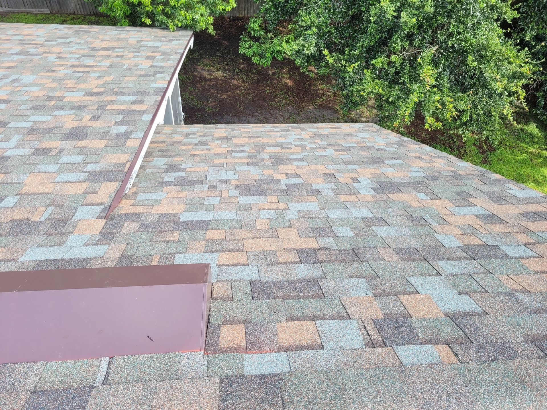 Close-up of a multi-colored shingled roof with a dark brown edge, with greenery visible in the background.