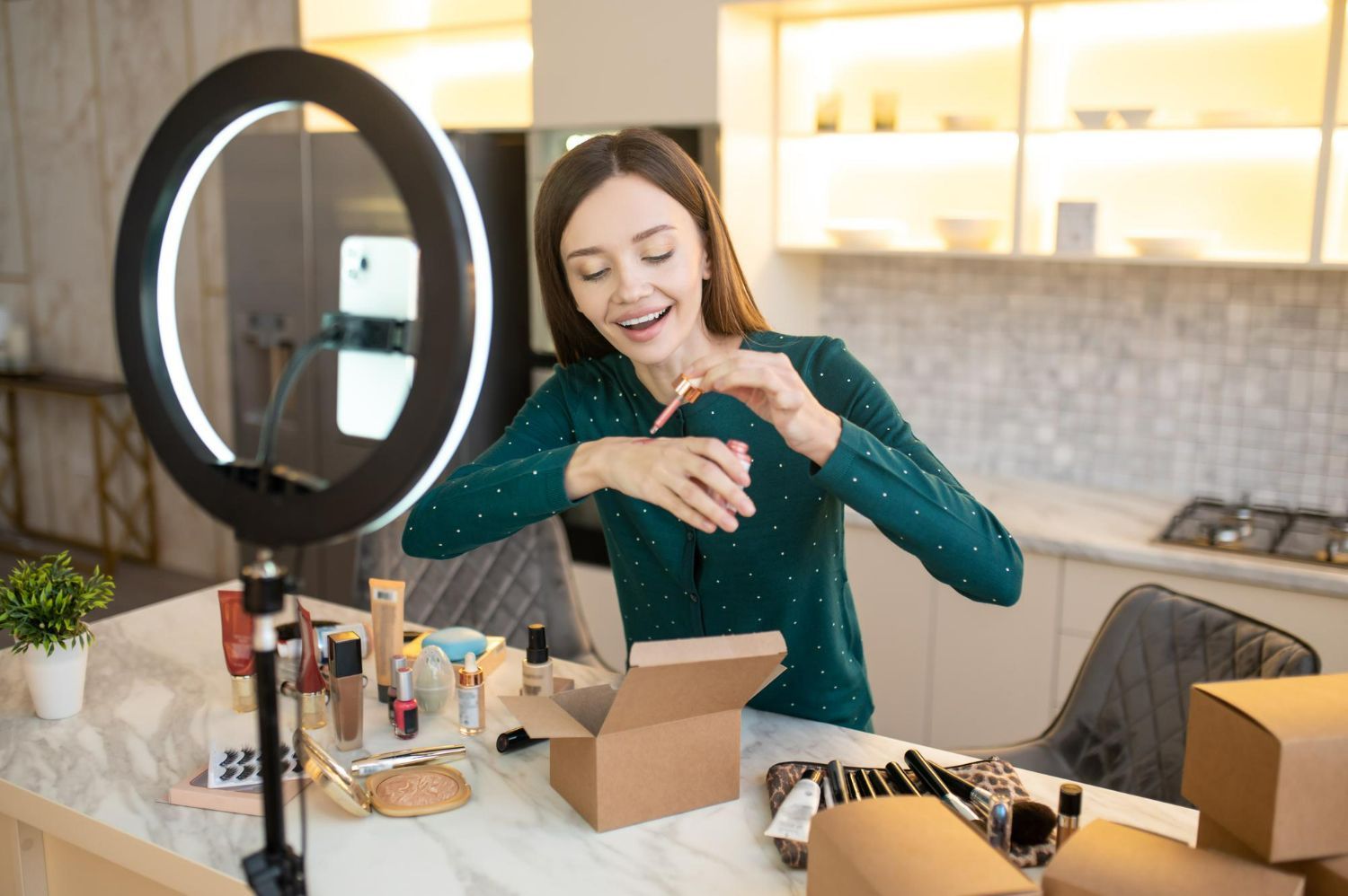 A woman is sitting at a table with a ring light and a box of makeup.