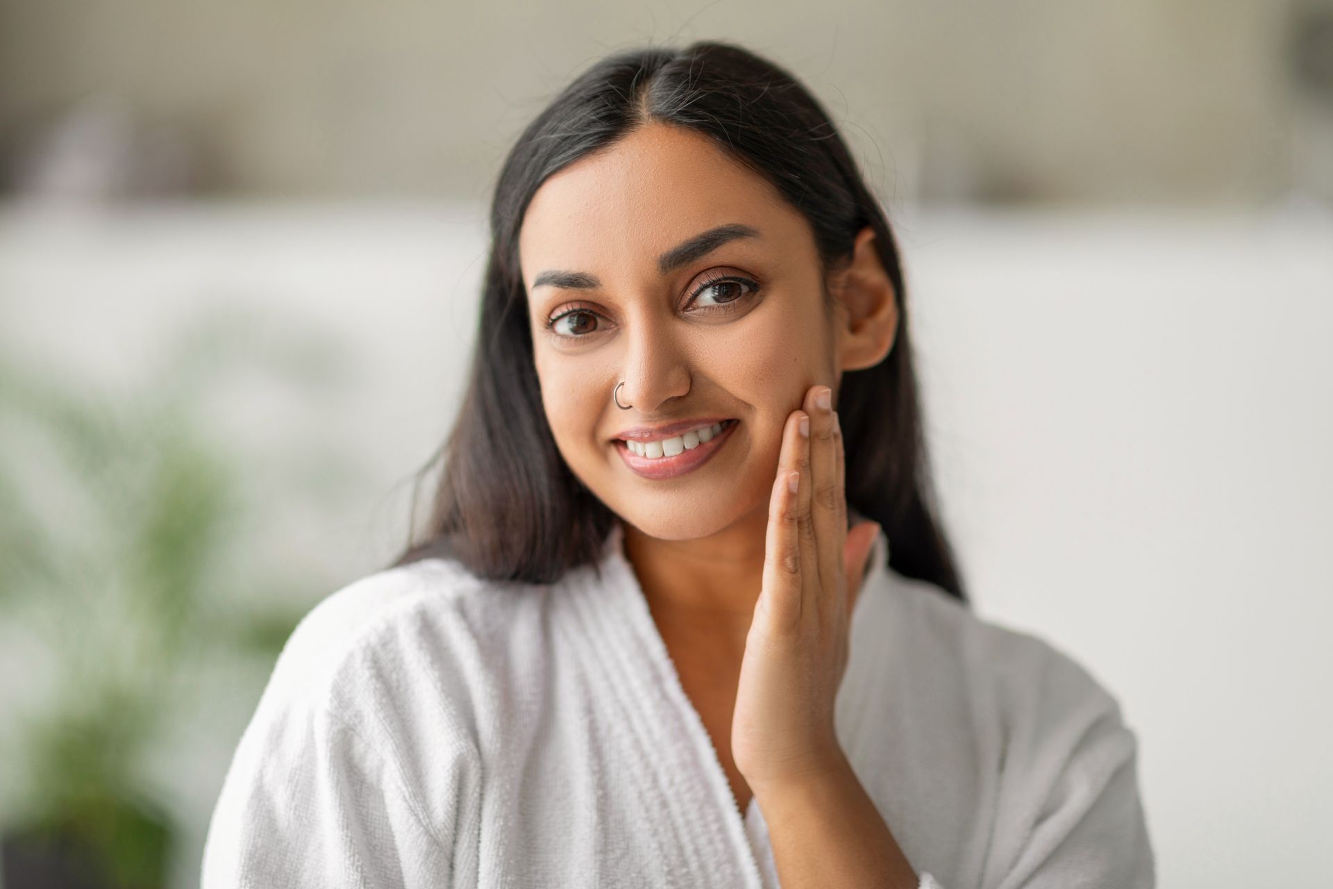 A woman in a bathrobe is touching her face and smiling.