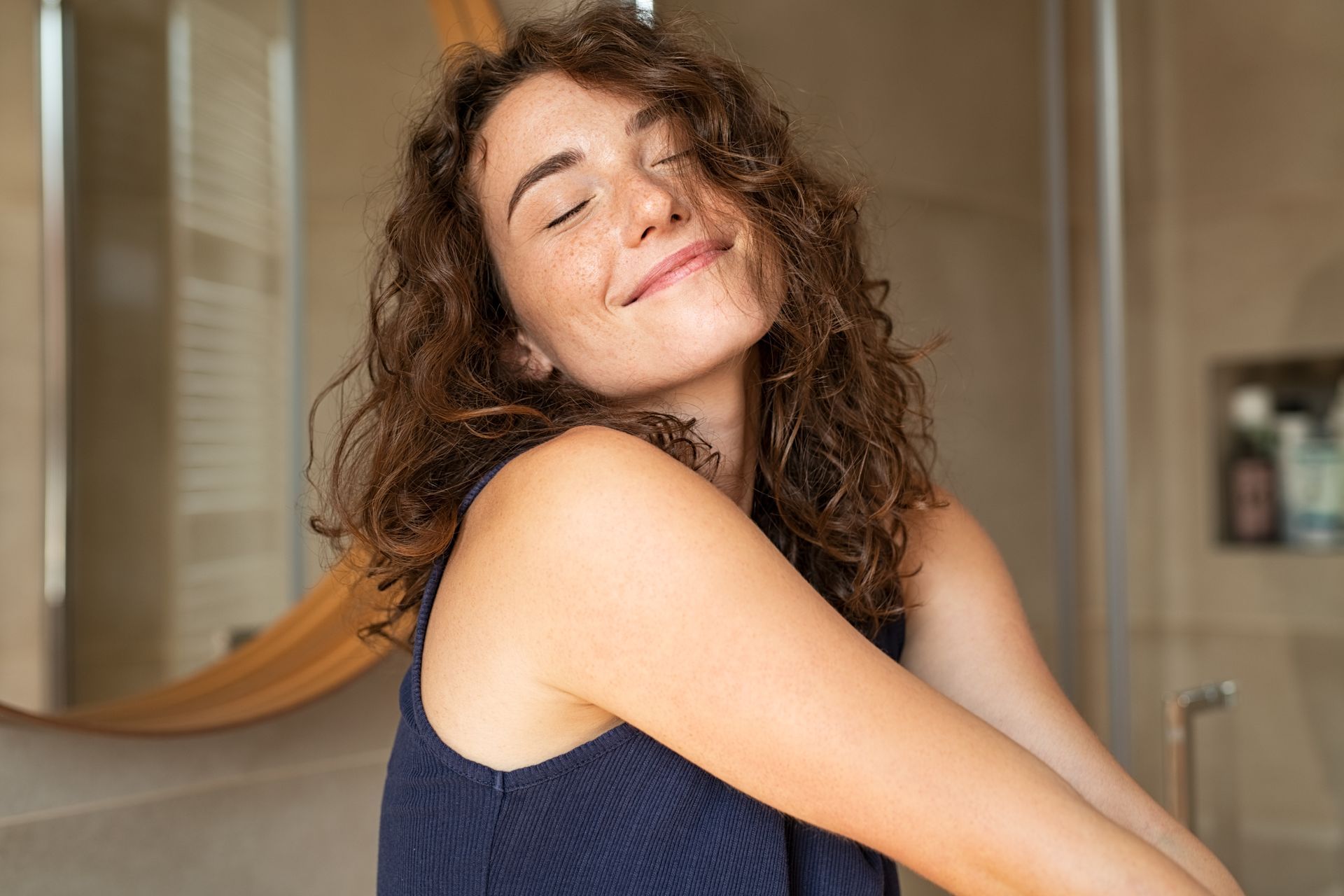 A woman with curly hair is smiling in a bathroom with her eyes closed.