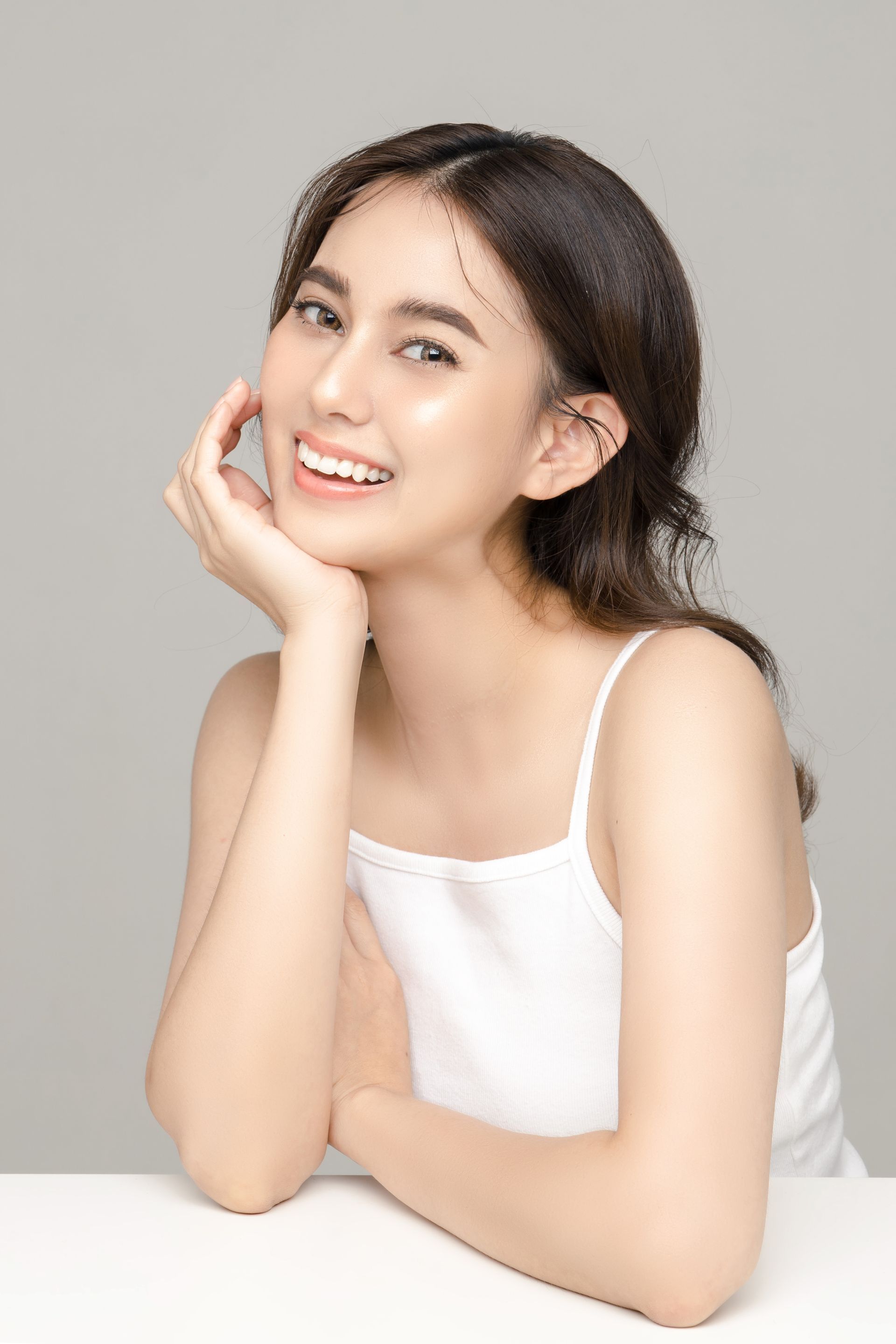 A woman in a white tank top is smiling with her hand on her face.