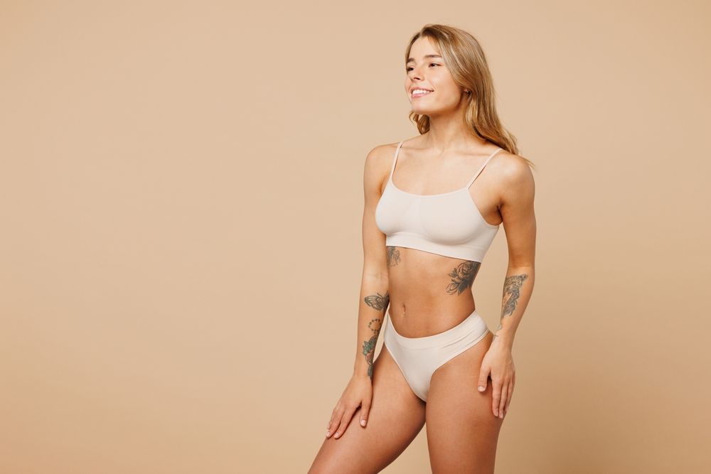 A woman in underwear with tattoos on her body is standing on a beige background.