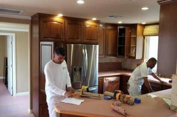 Two men are painting a kitchen with wooden cabinets and stainless steel appliances.