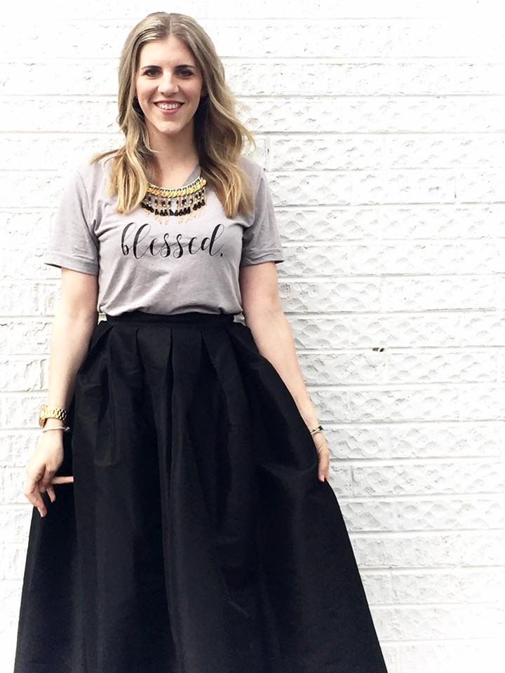 A woman wearing a gray t-shirt and a black skirt is standing in front of a white brick wall.