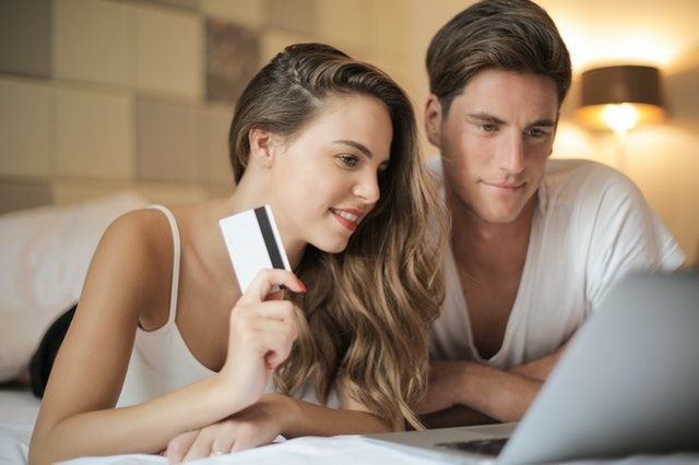 A man and a woman are laying on a bed looking at a laptop . the woman is holding a credit card.