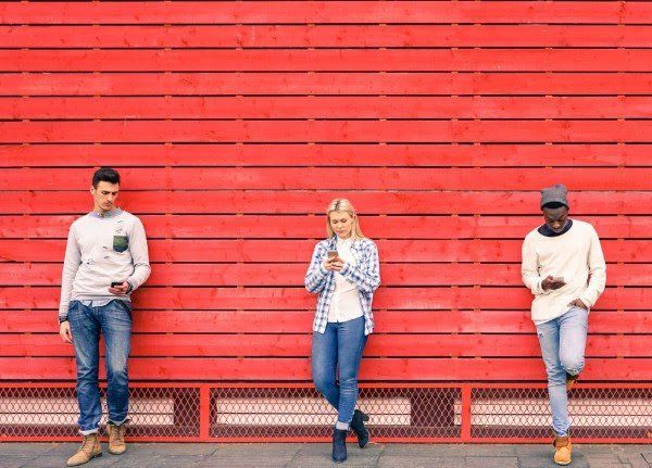 Three people are standing in front of a red wall looking at their phones.