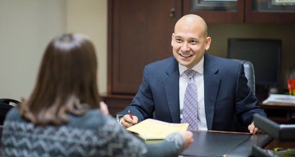 A man in a suit and tie is sitting at a desk talking to a woman.