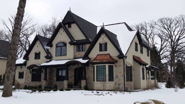 A large white house with a black roof is surrounded by snow and trees.