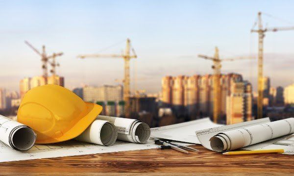 A hard hat and blueprints are on a wooden table with a construction site in the background.