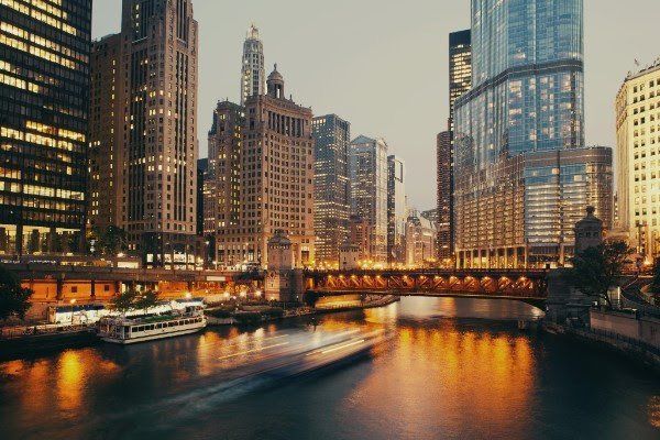 A boat is floating down a river in front of a city skyline at night.