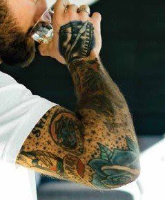 A man with a lot of tattoos on his arms is drinking from a glass.