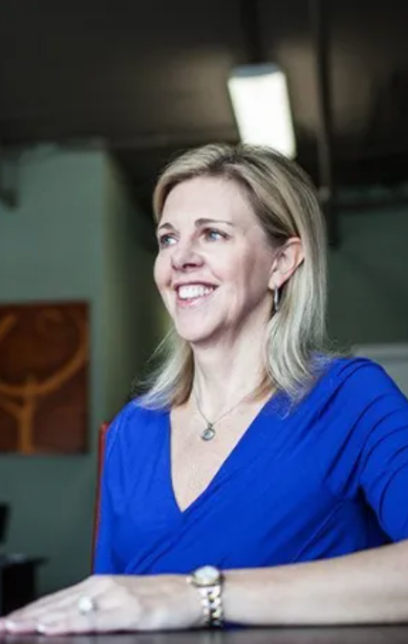A woman in a blue shirt is sitting at a table and smiling.
