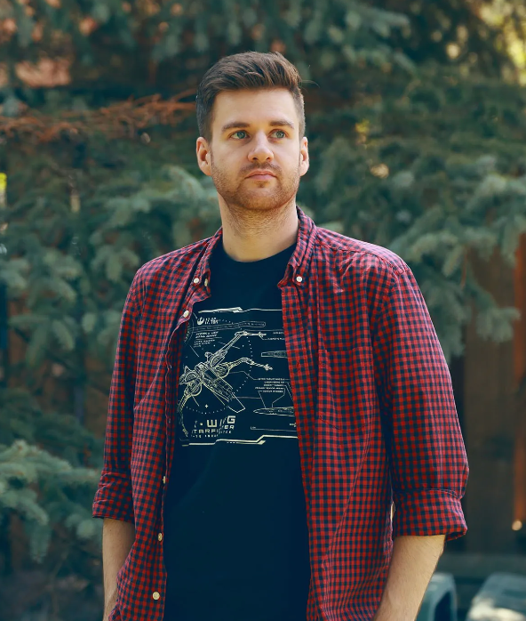 A man wearing a plaid shirt and a black t-shirt is standing with his hands in his pockets.