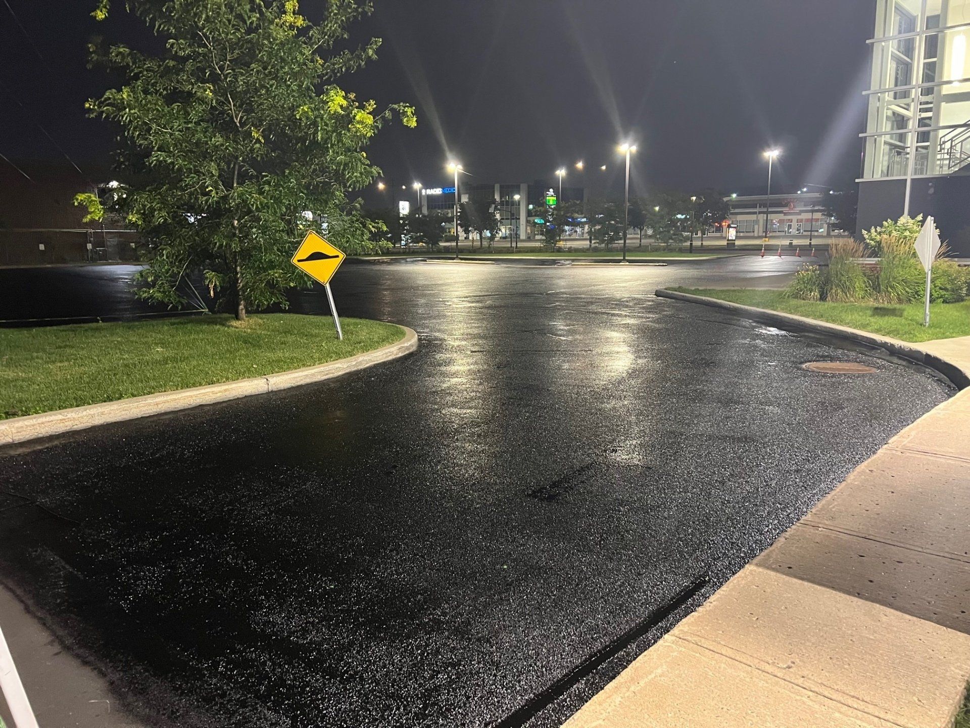 Une route asphaltée noire et humide serpente devant une zone herbeuse et un bâtiment ; panneau de signalisation jaune ; nuit.