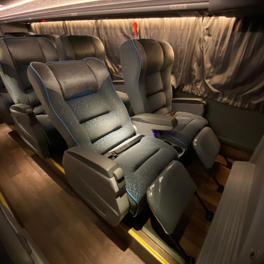 Assentos de ônibus cinzas e reclináveis, com apoios para os pés, em uma cabine mal iluminada.