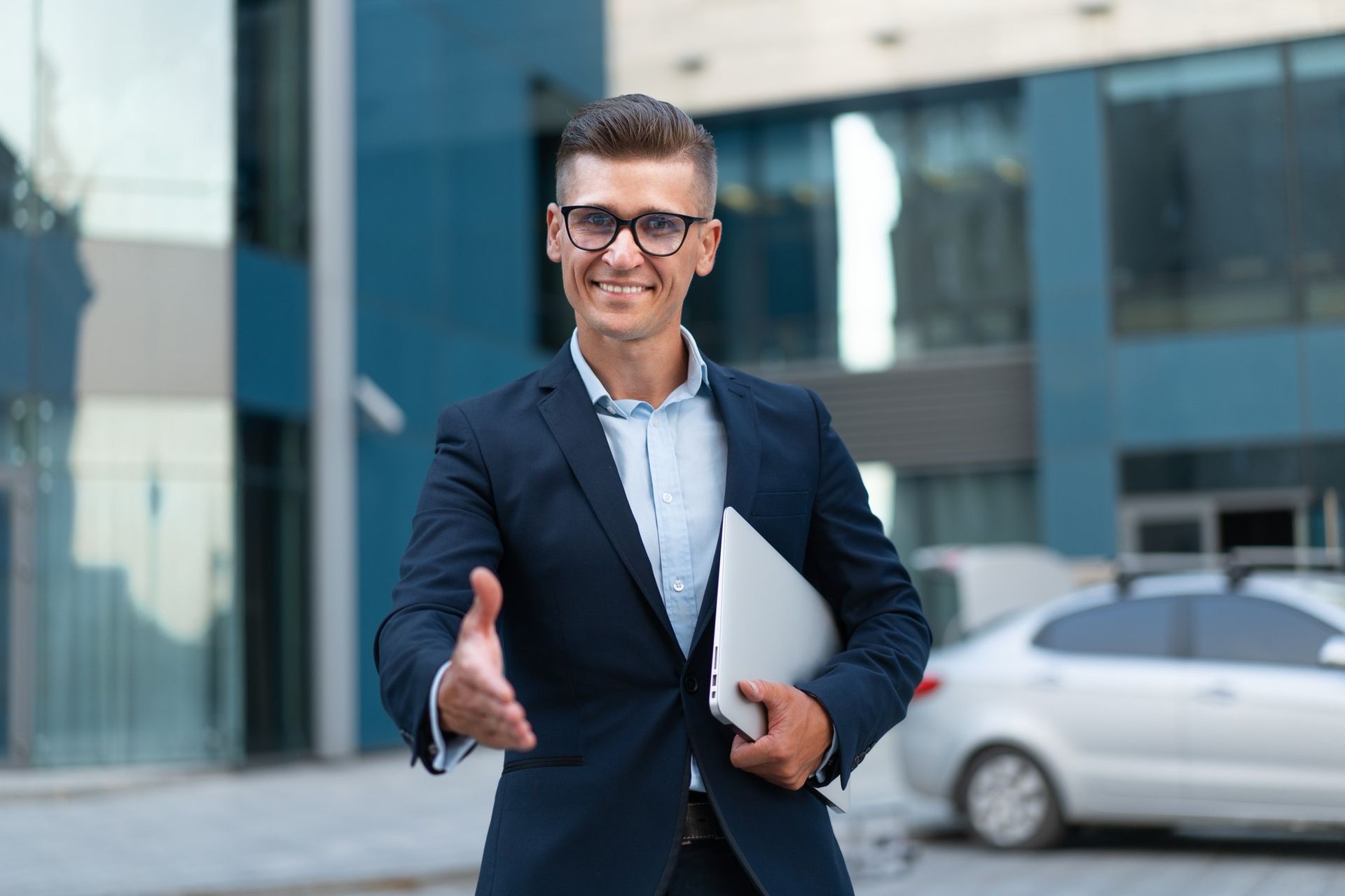Smiling businessman in a navy suit shaking hands outside a modern office building, holding a folder