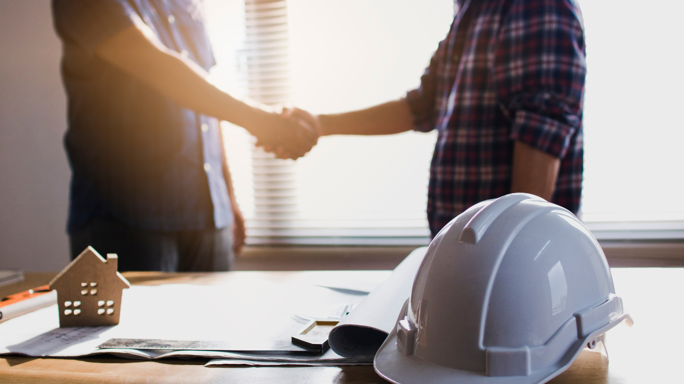 Two men are shaking hands over a table with a hard hat and a model house.