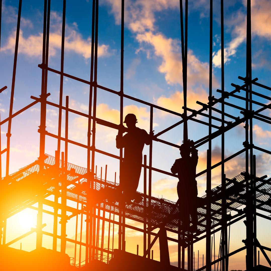 Two construction workers are standing on scaffolding at a construction site at sunset.