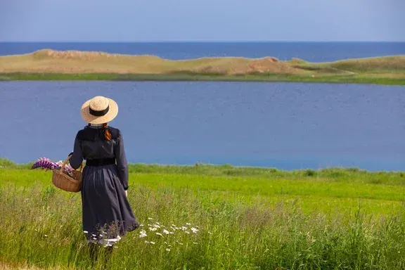 A woman in a straw hat is standing in a field looking at the ocean.