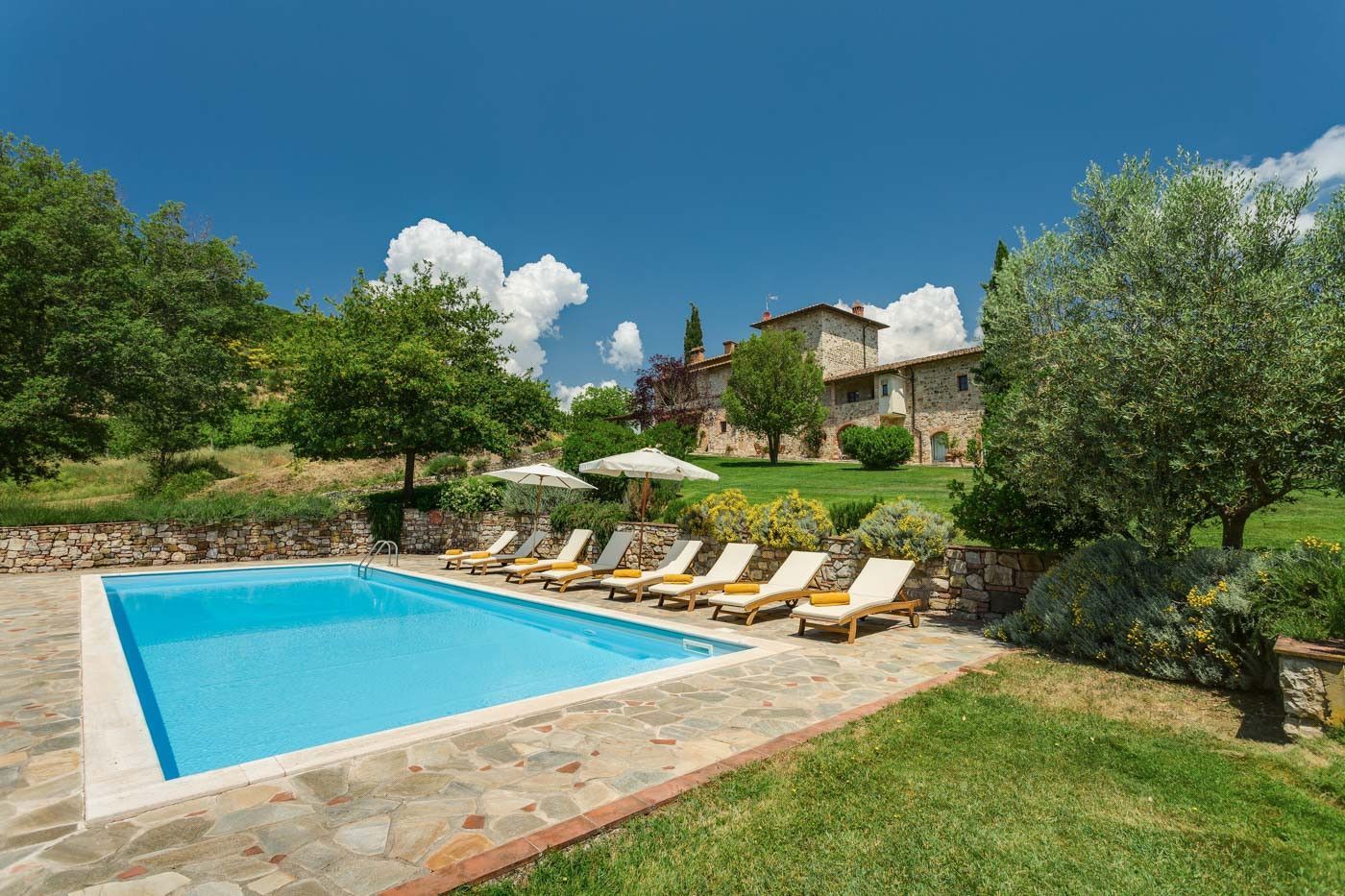 A large swimming pool surrounded by chairs and umbrellas in front of a villa in Tuscany.