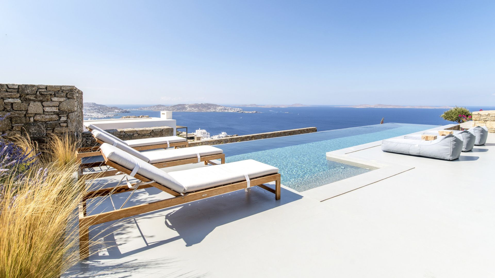 A row of lounge chairs sitting next to a swimming pool overlooking the ocean at villa in Mykonos, Greece.