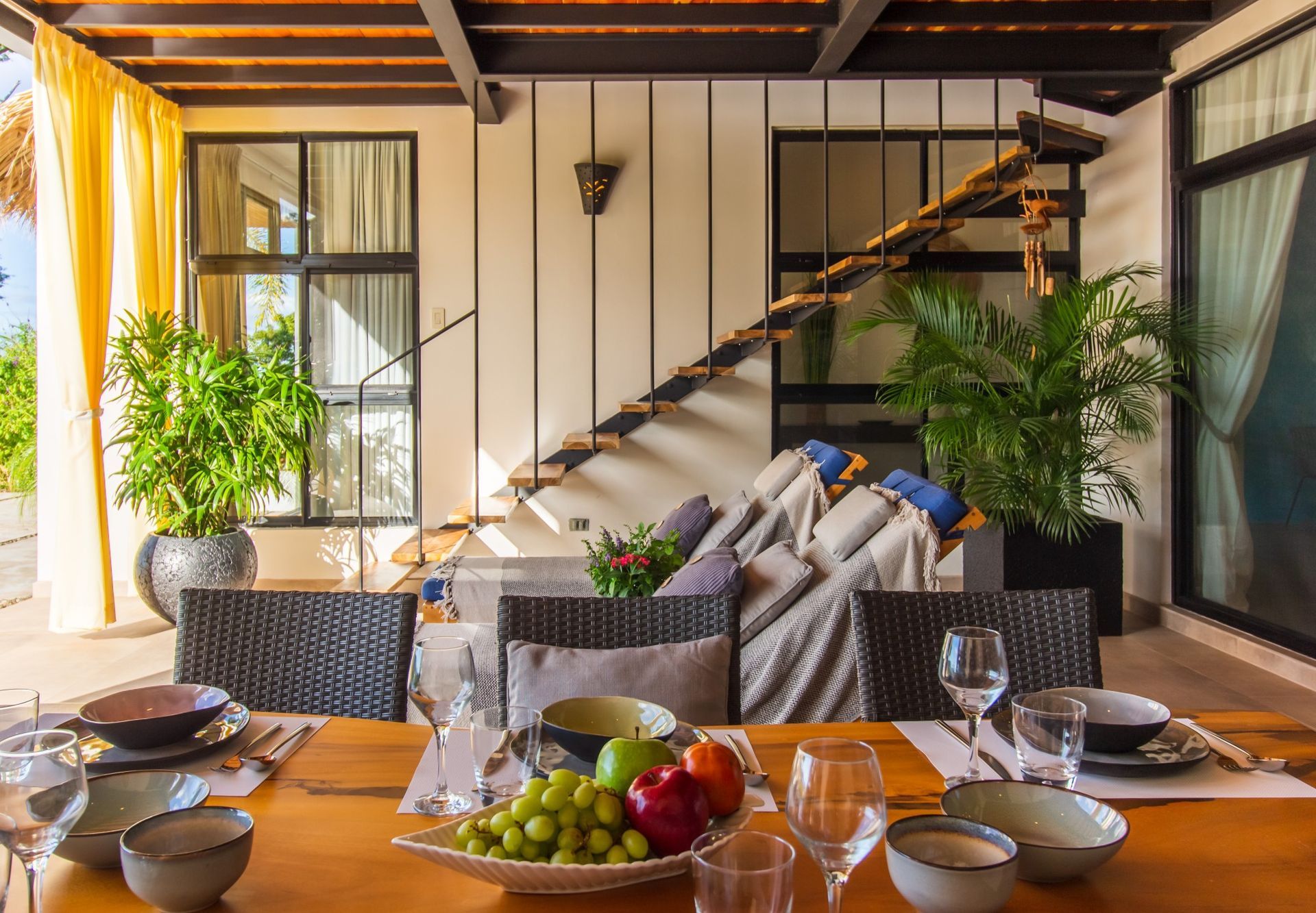 A dining table with plates , bowls , glasses and fruit on it in a living room with a staircase at villa in Costa Rica.