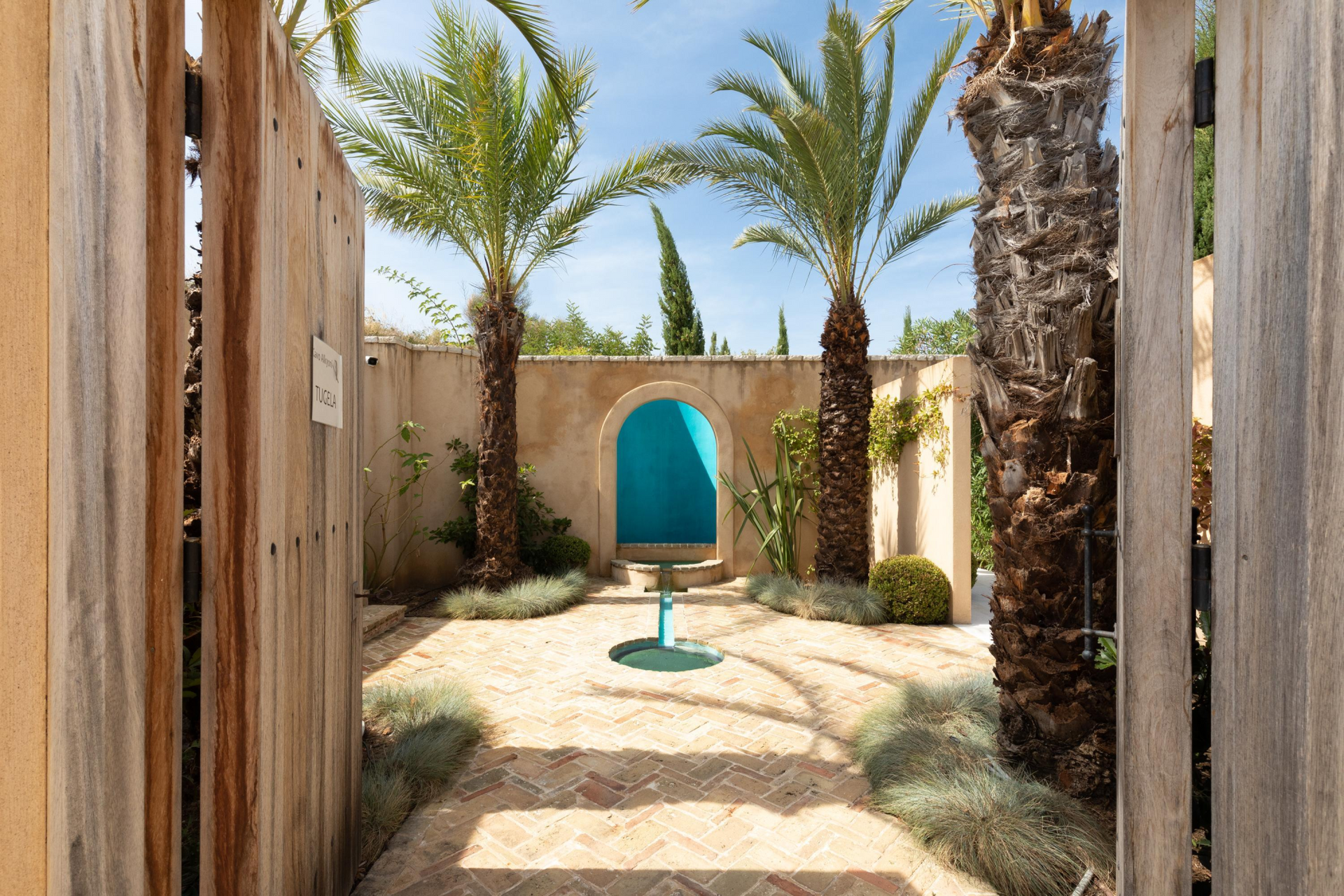 A wooden gate leads to a patio with palm trees and a blue chair at villa in Corfu, Greece.