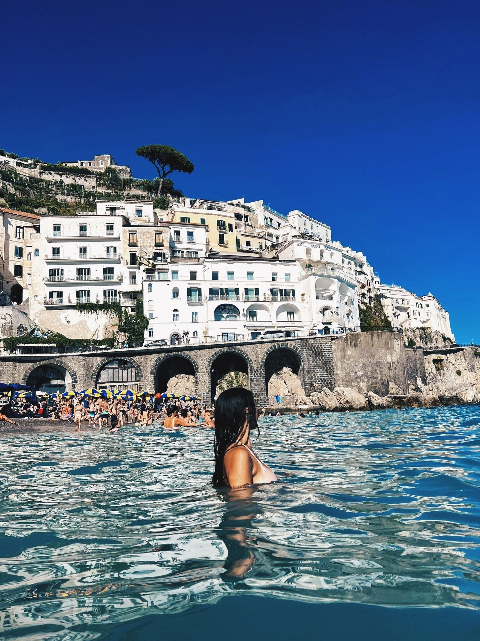 A woman is swimming in the ocean with the Amalfi Coast in the background in Italy.