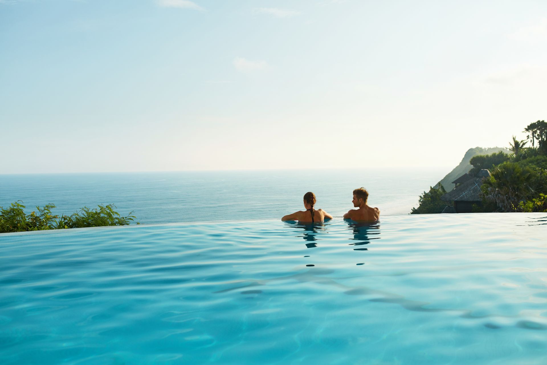 Two people are swimming in an infinity pool overlooking the ocean.