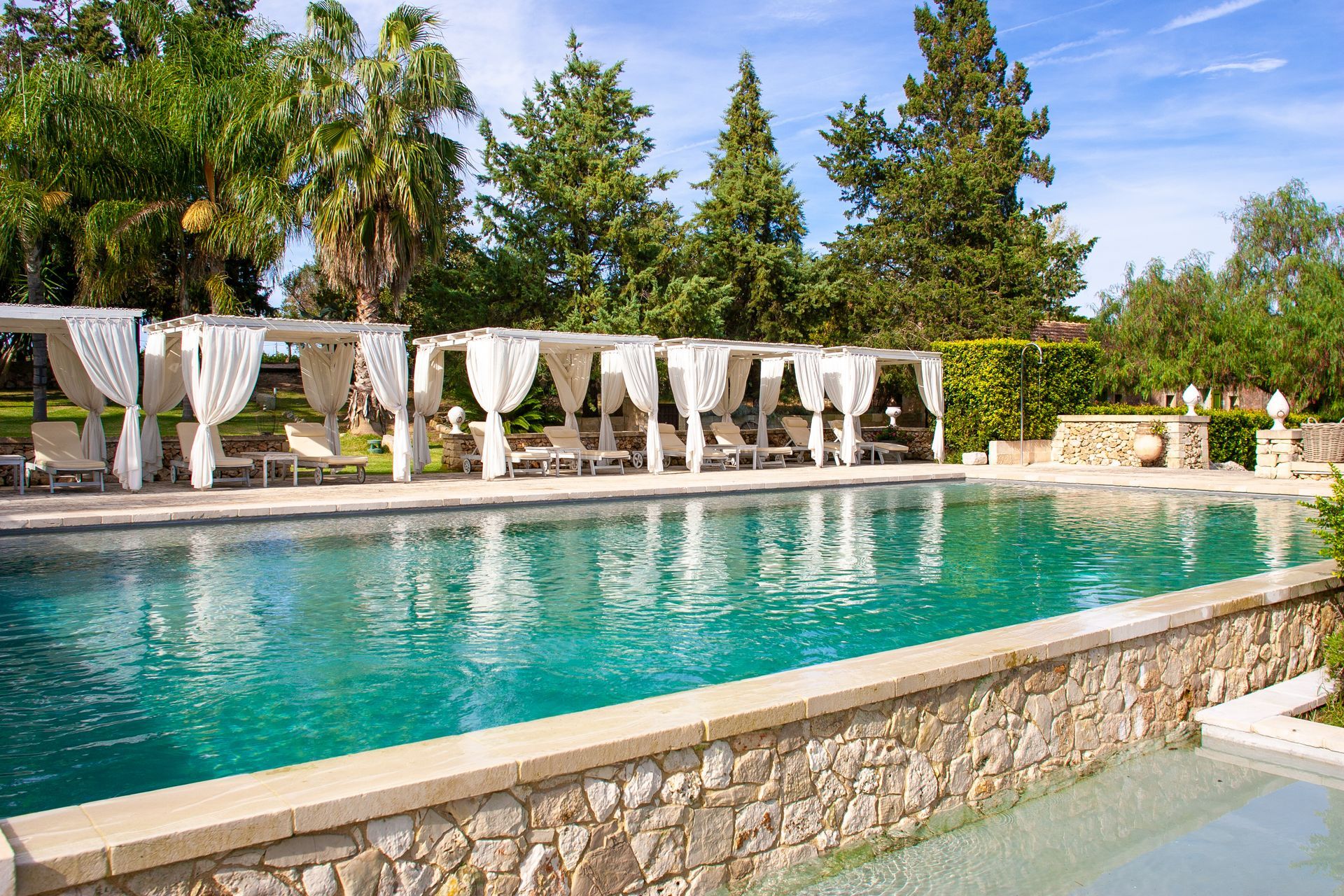 A large swimming pool surrounded by trees and a stone wall at Naturalis Bio Resort & Spa in Puglia, Italy.