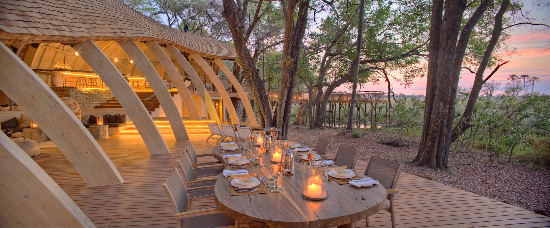 There is a long table with candles on it in the middle of the woods at a camp in Africa.