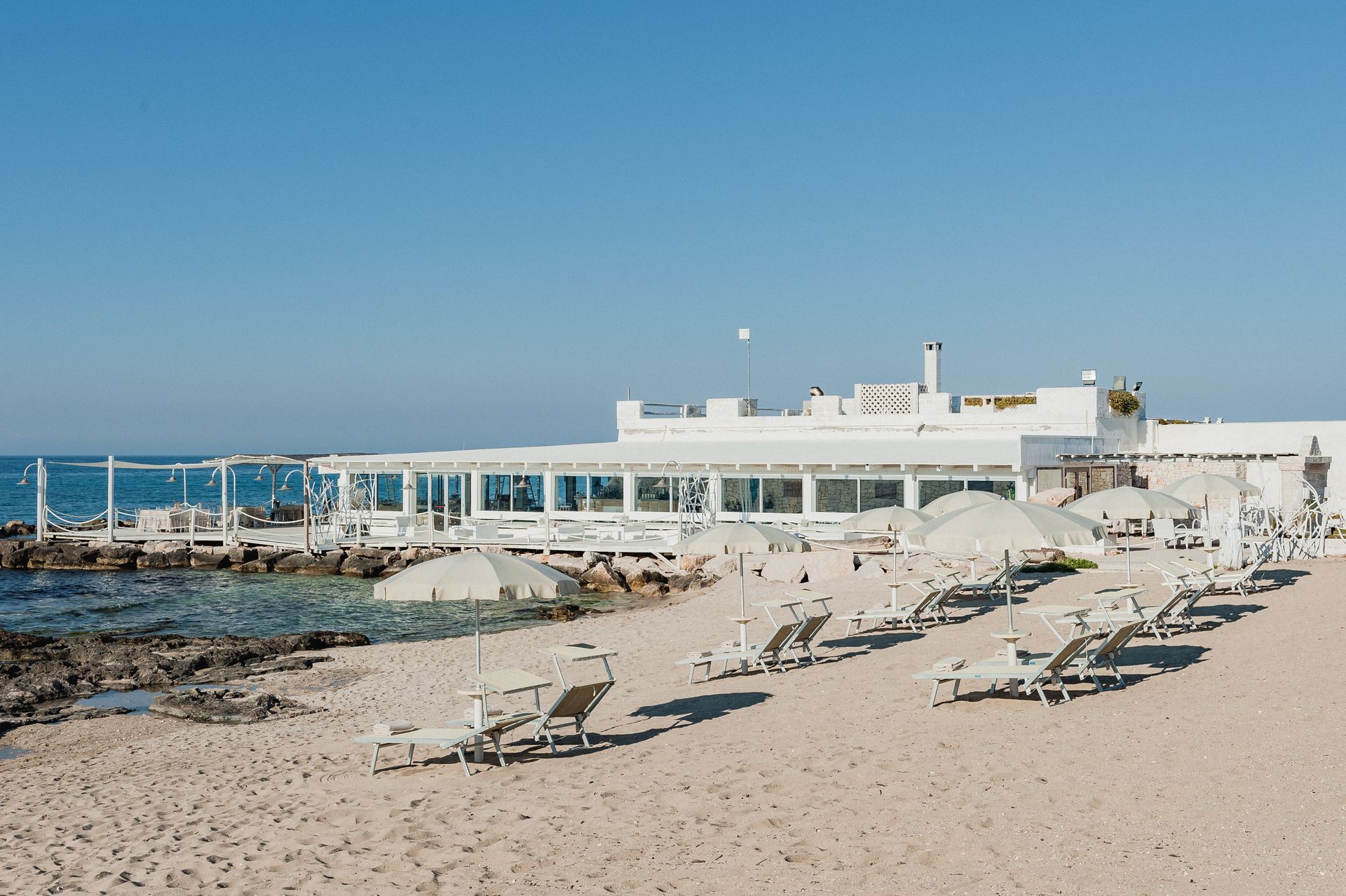 A beach with chairs and umbrellas in front of a white building at La Peschiera Hotel in Puglia, Italy.