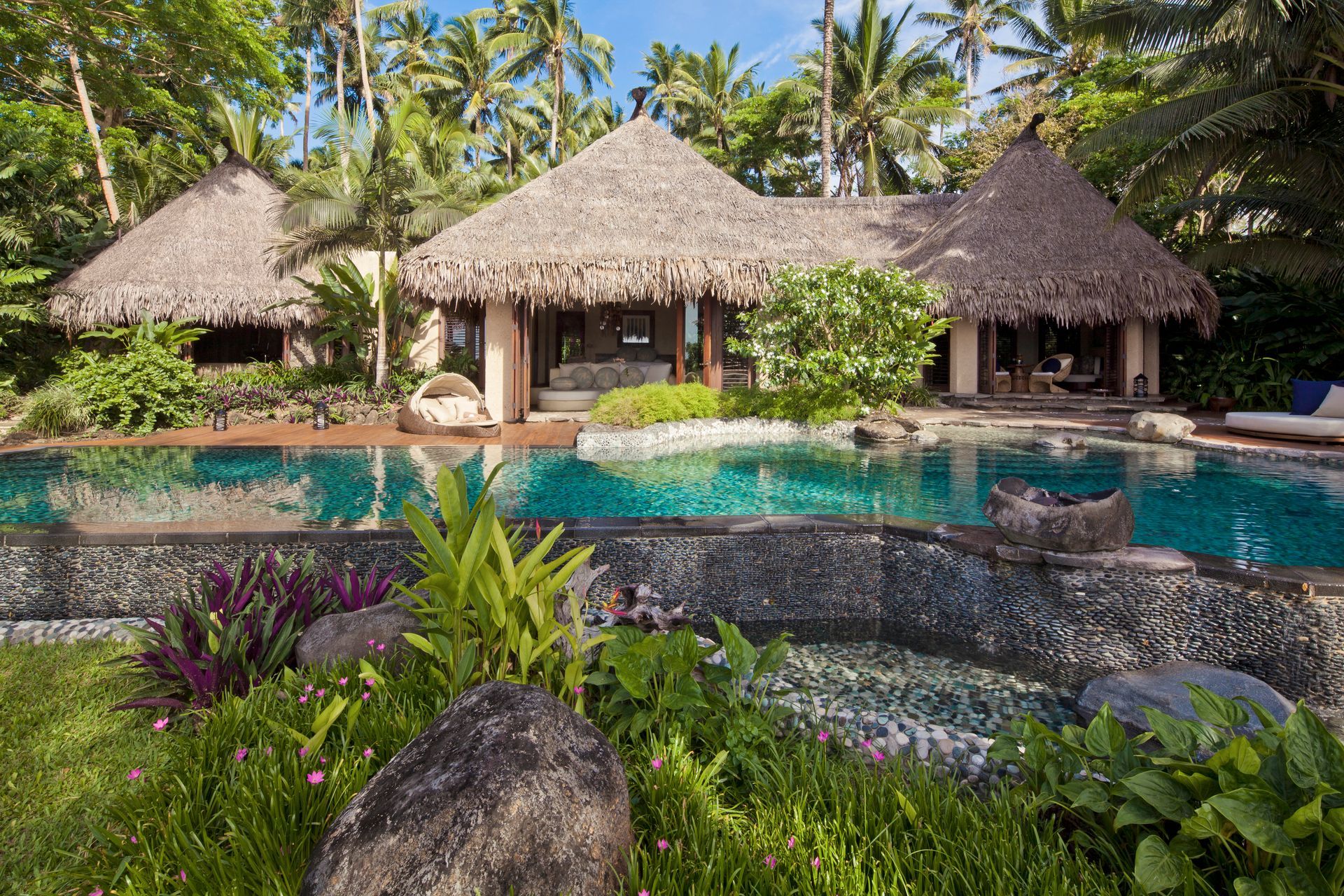 There is a large swimming pool in front of a villa with thatched roofs in Fiji.