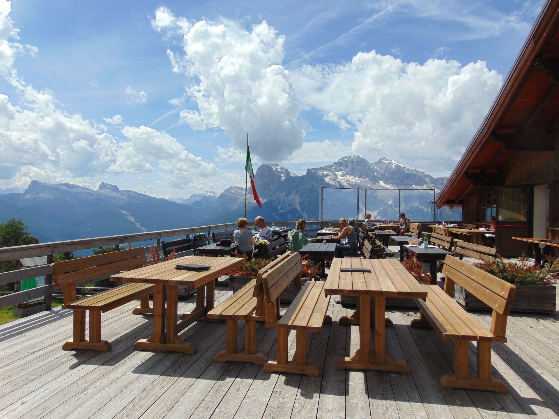 Wooden tables and benches on a deck overlook mountains, Italian flag. Several people are seated. Sunny day.