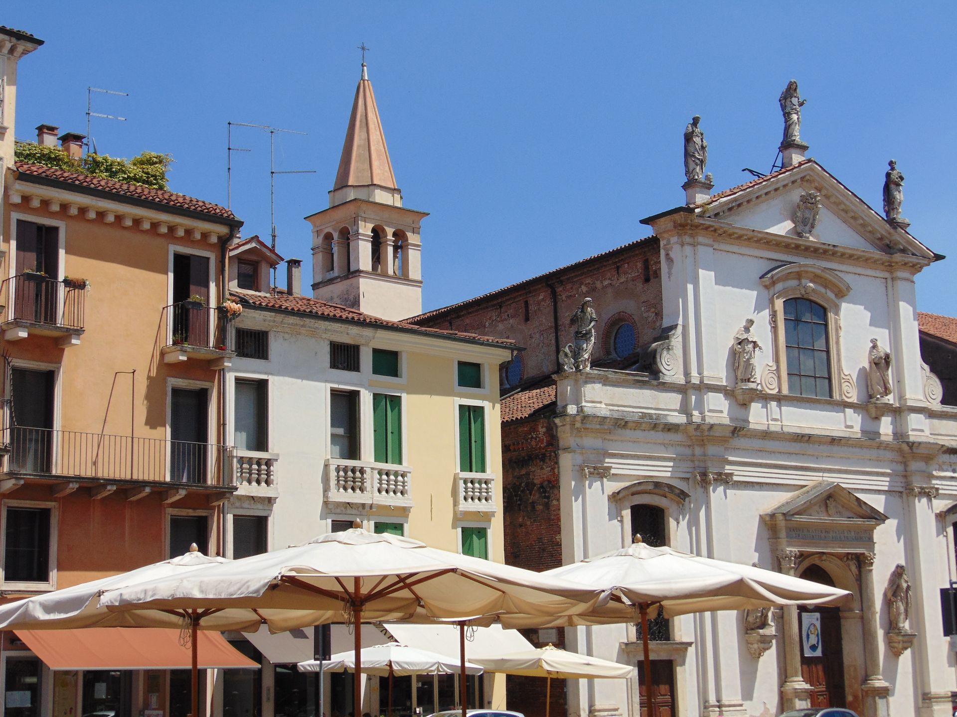 Buildings in a sunny square with a church steeple and umbrellas covering outdoor seating.