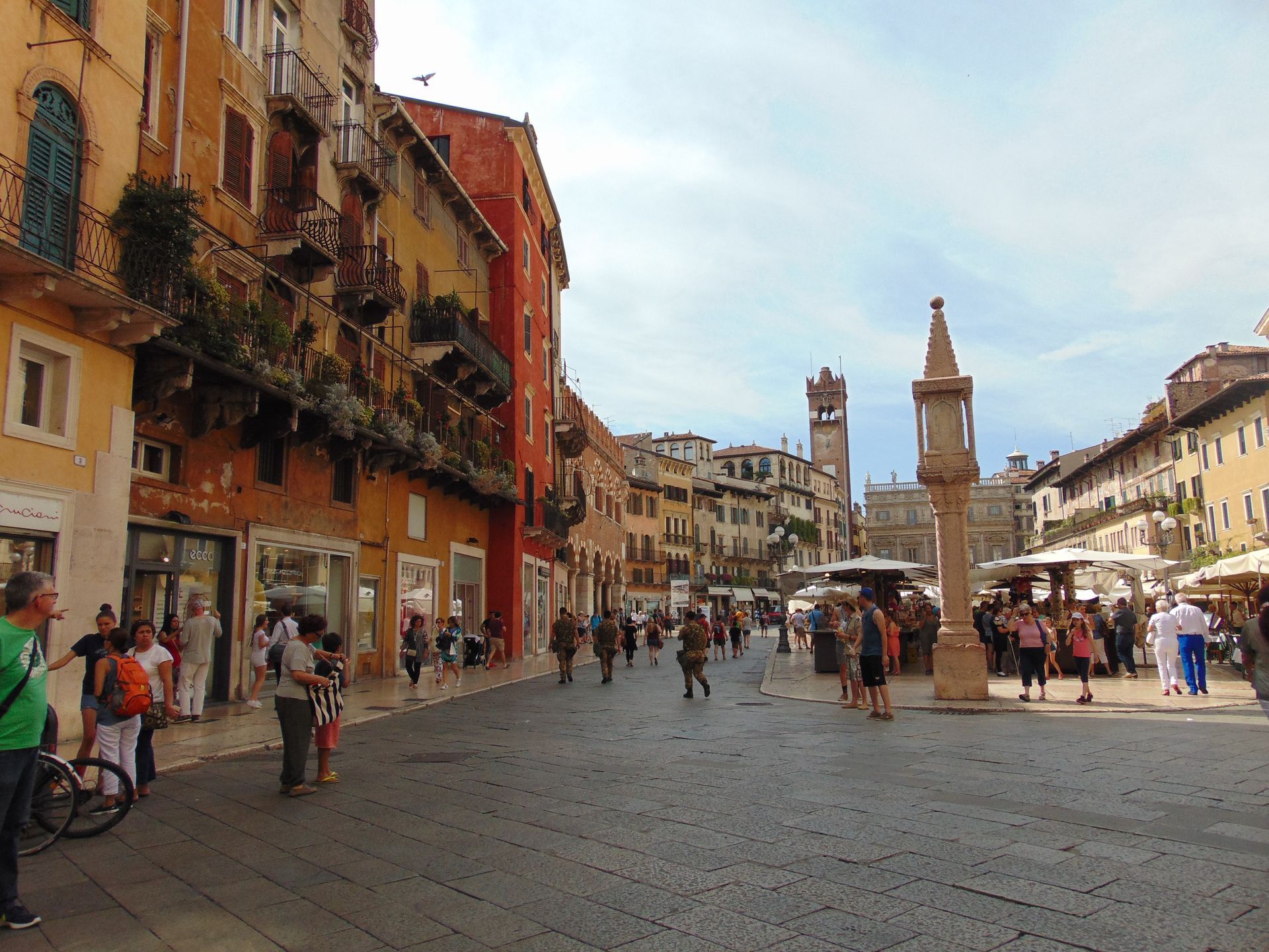 Cobblestone square in Verona, Italy, with historic buildings, pedestrians, and a tall monument.