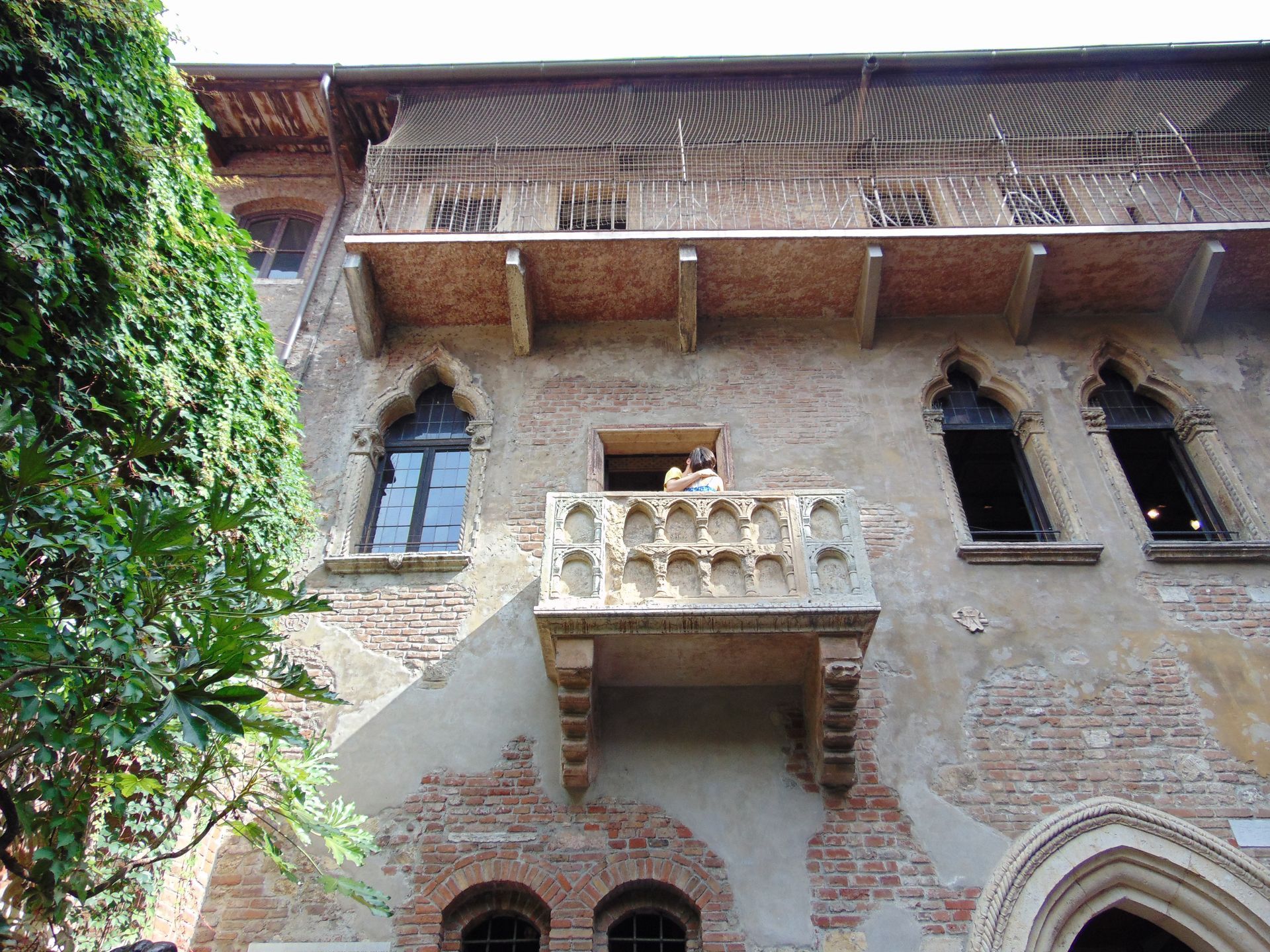 Juliet's balcony on a weathered brick building in Verona, Italy, with arched windows and a partial view of ivy.