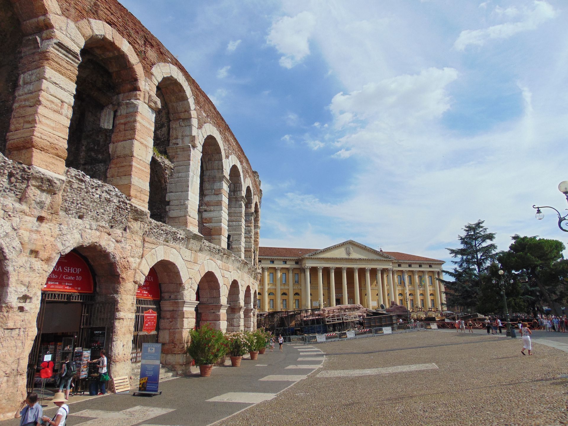 Verona Arena exterior, stone arches, people walking, large building in background, blue sky.