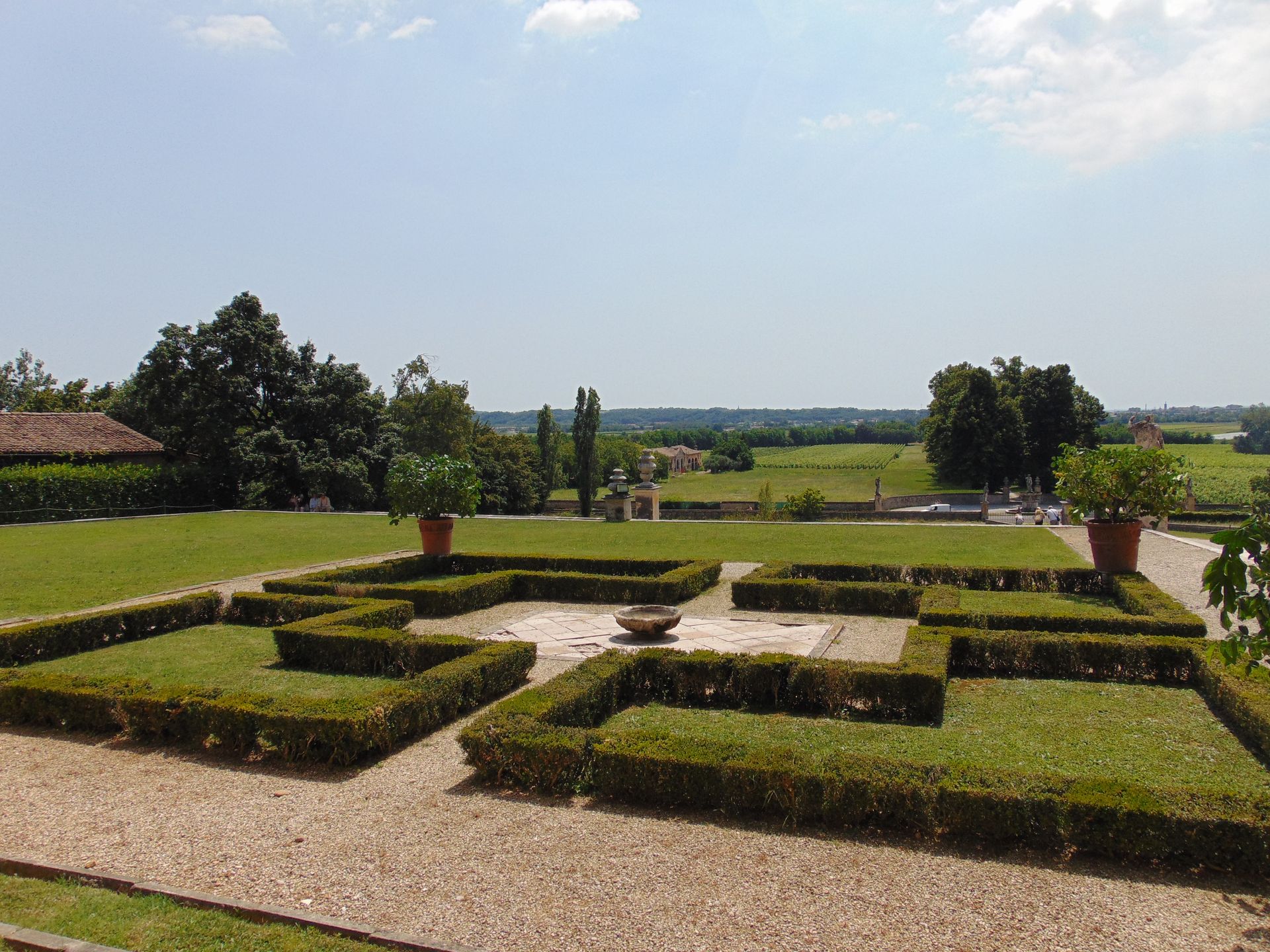 Formal garden with trimmed hedges, pots, and distant landscape under a blue sky.