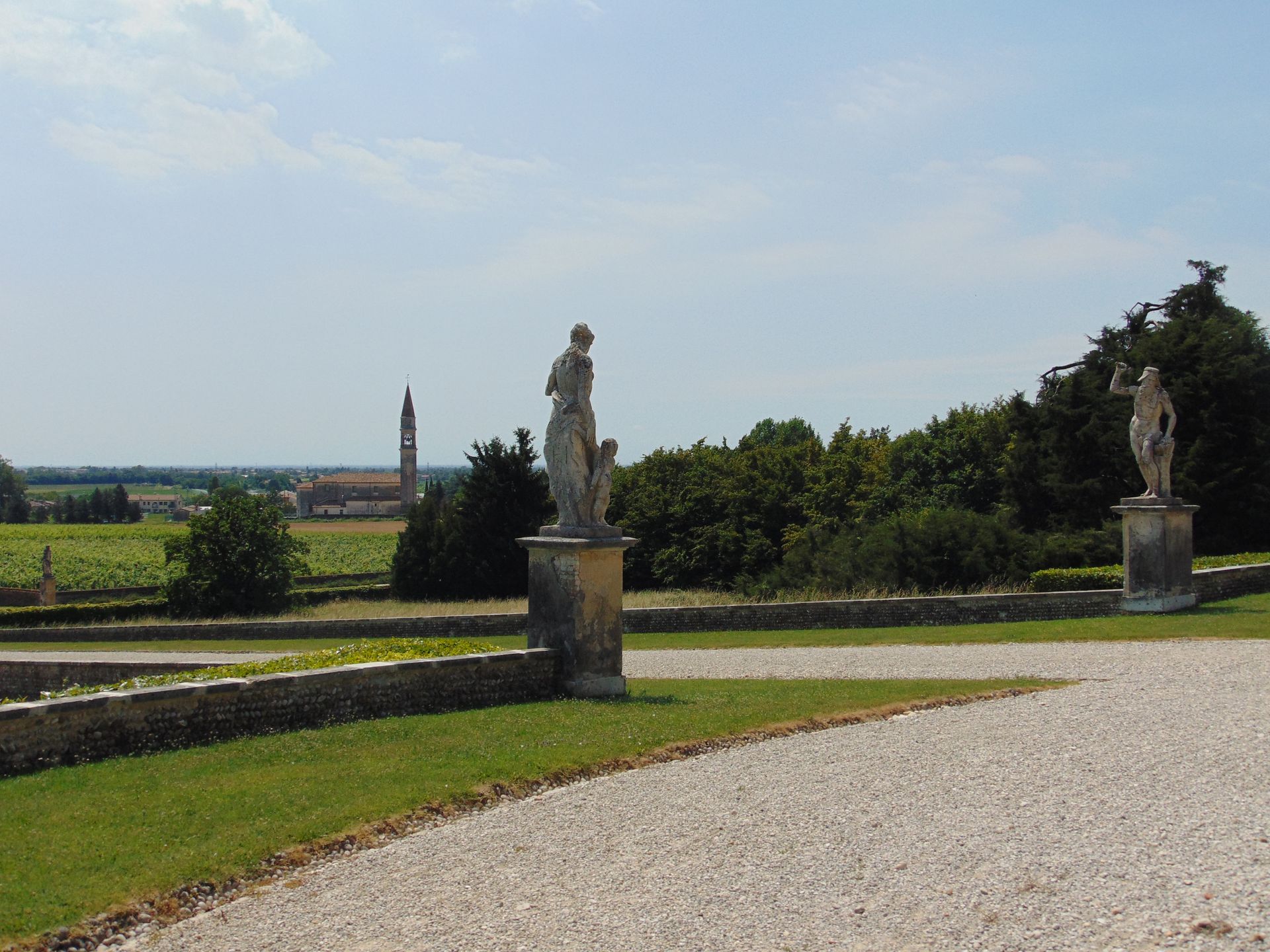 Stone statues in a formal garden overlook a landscape with trees, fields, and a distant tower.