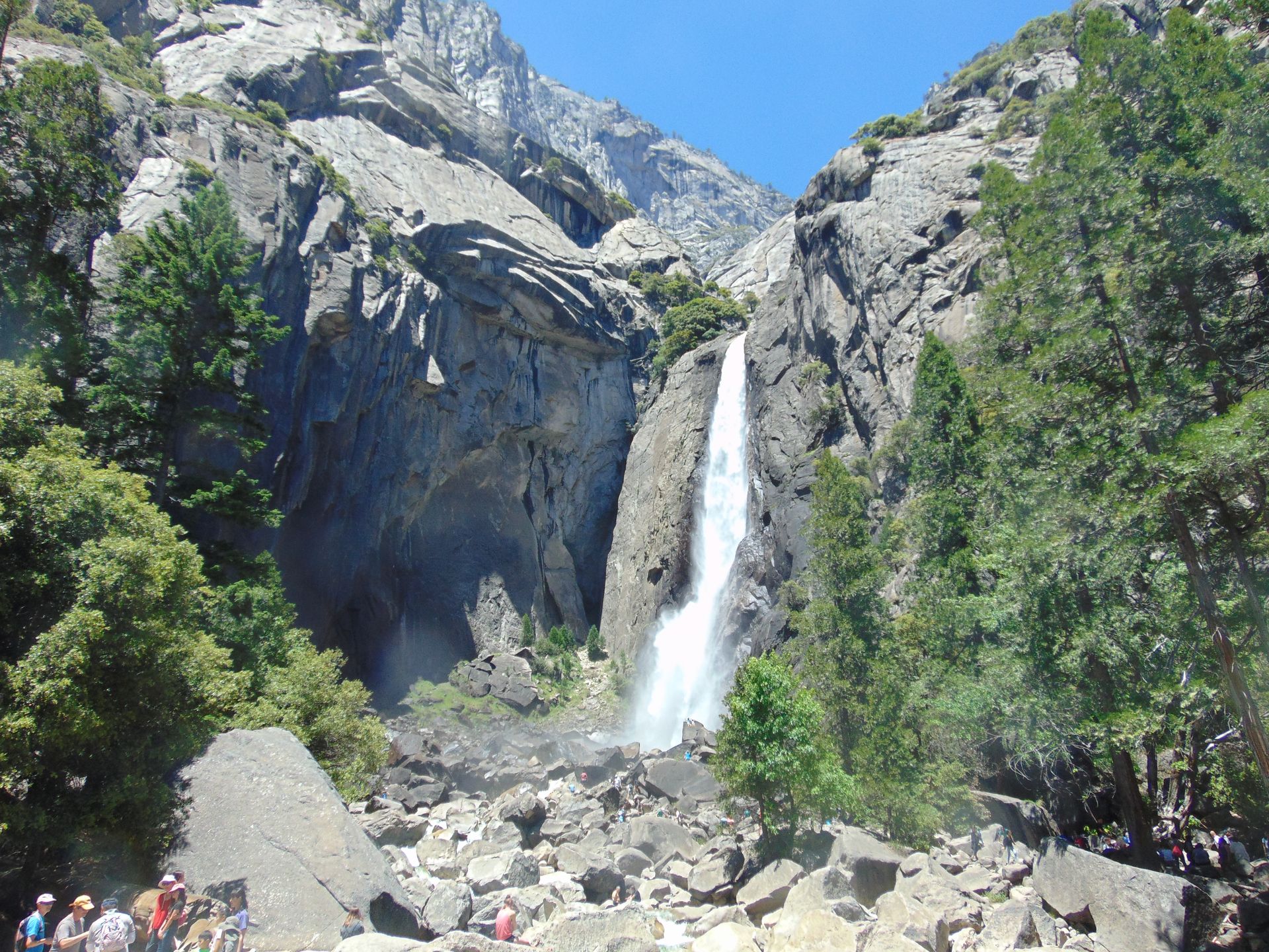 Yosemite waterfall cascading down a granite mountain, framed by green trees against a bright blue sky.