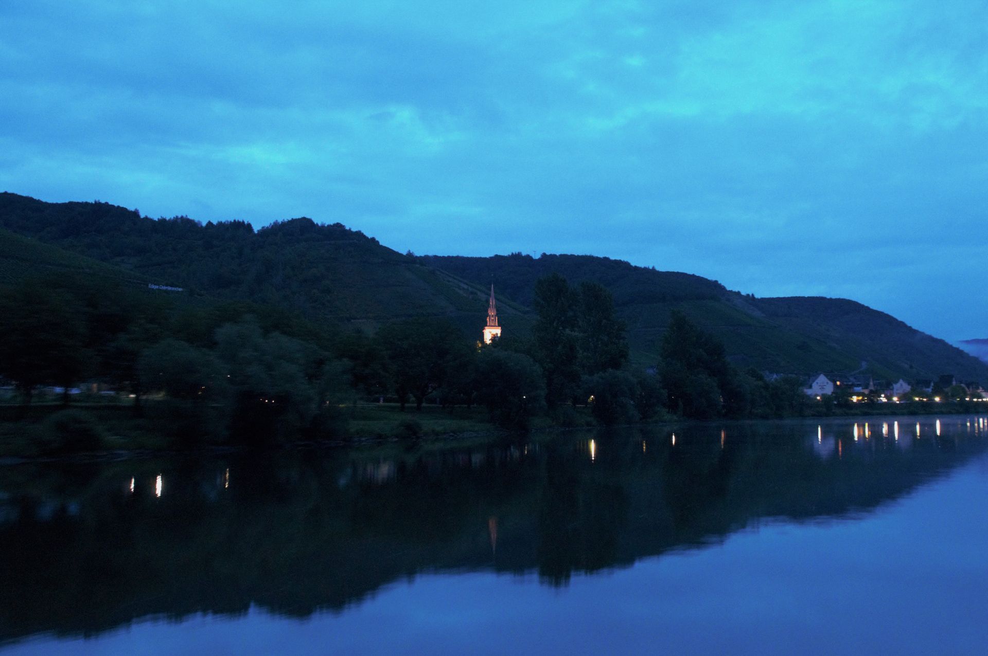 River at dusk, mountain and town on the banks. Church spire and lights reflected in water. Blue tones.