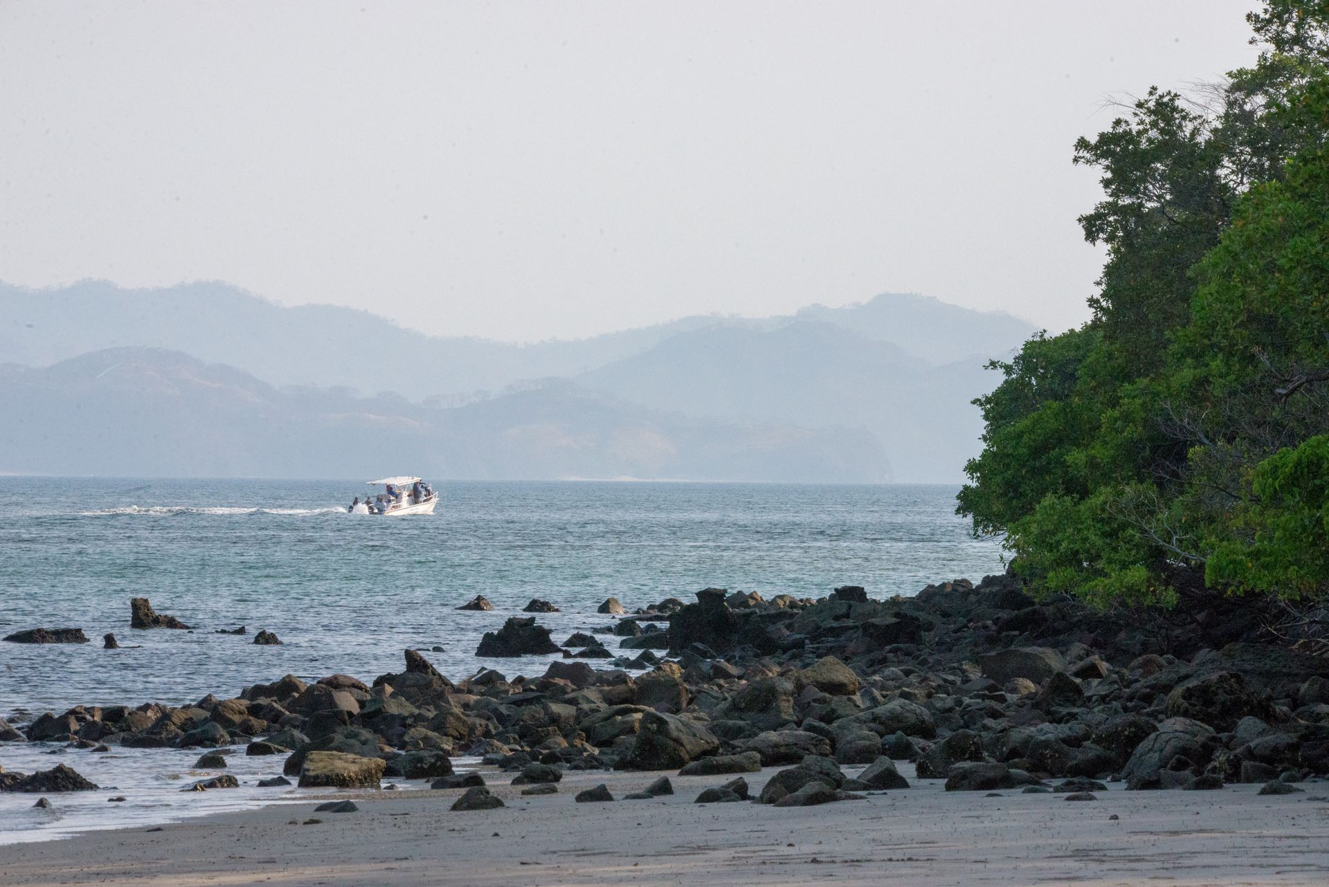 Rocky beach with water, mountains in the distance, and a boat. Trees are on the right.