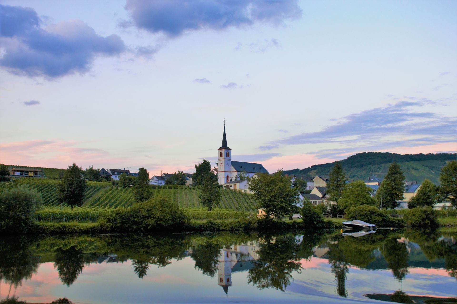 Church steeple reflected in calm water, overlooking a village with vineyards under a colorful sky.