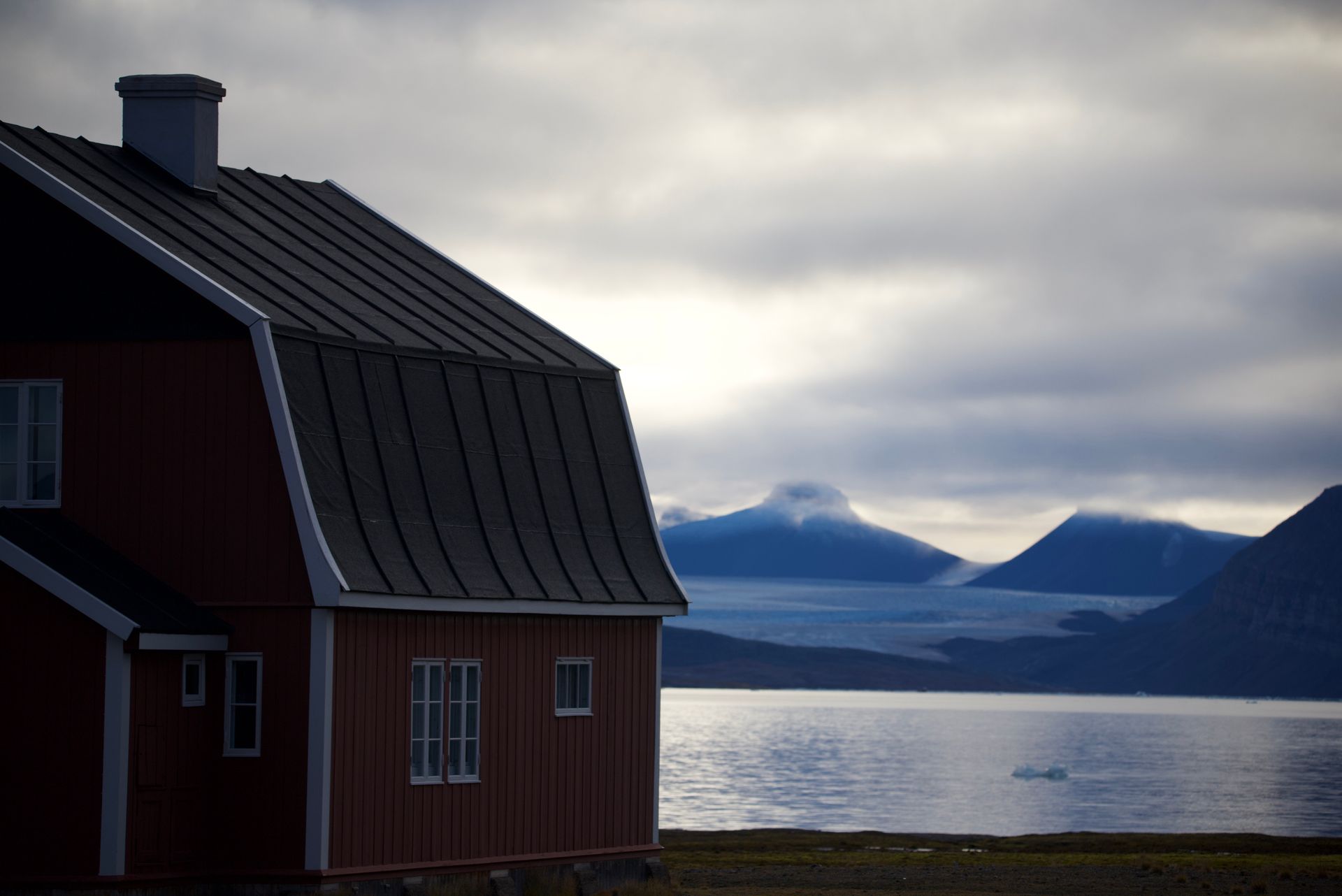 Red house with a dark roof near a body of water and mountains under a cloudy sky.