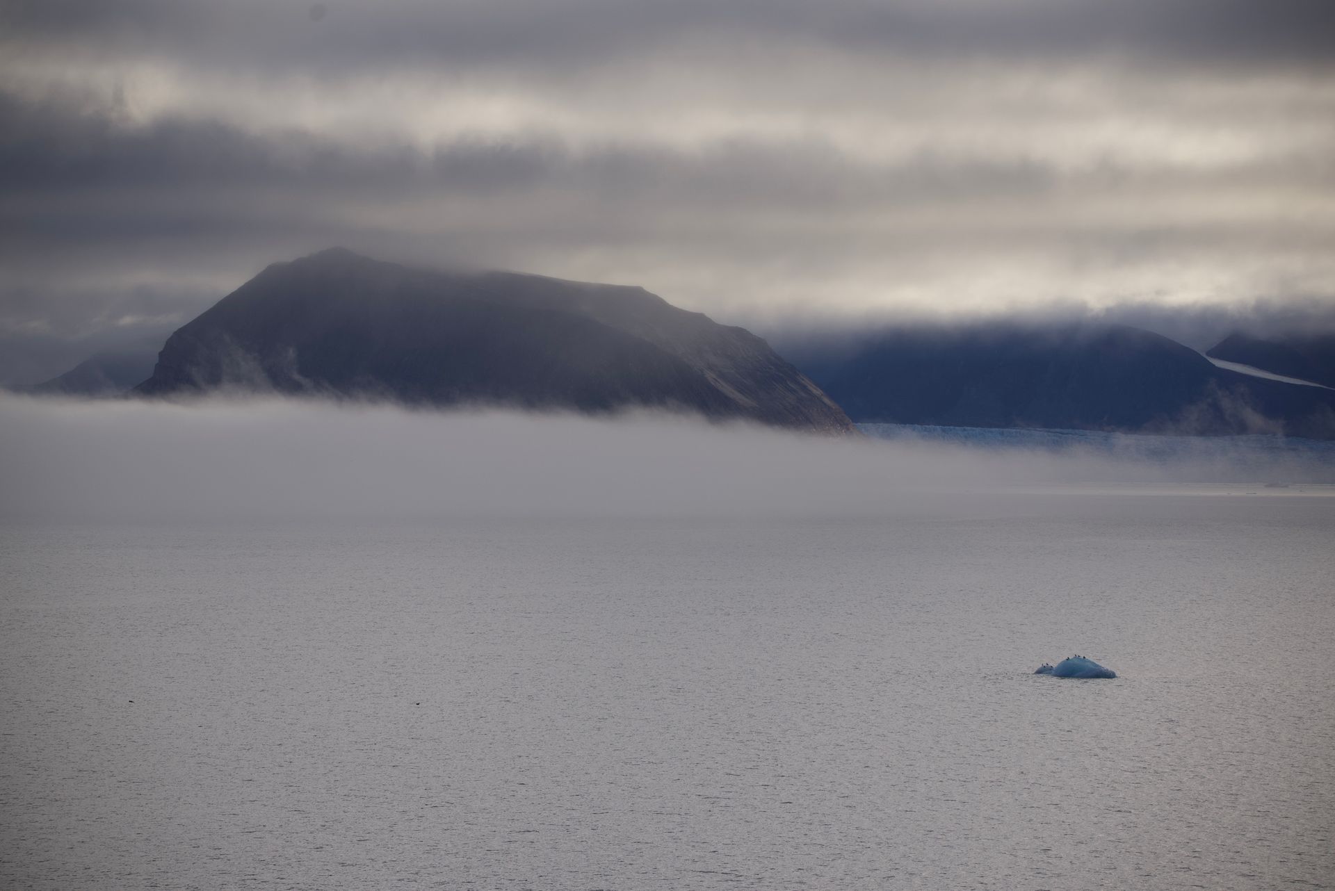 Foggy Arctic landscape with a mountain, low clouds, and a small iceberg.