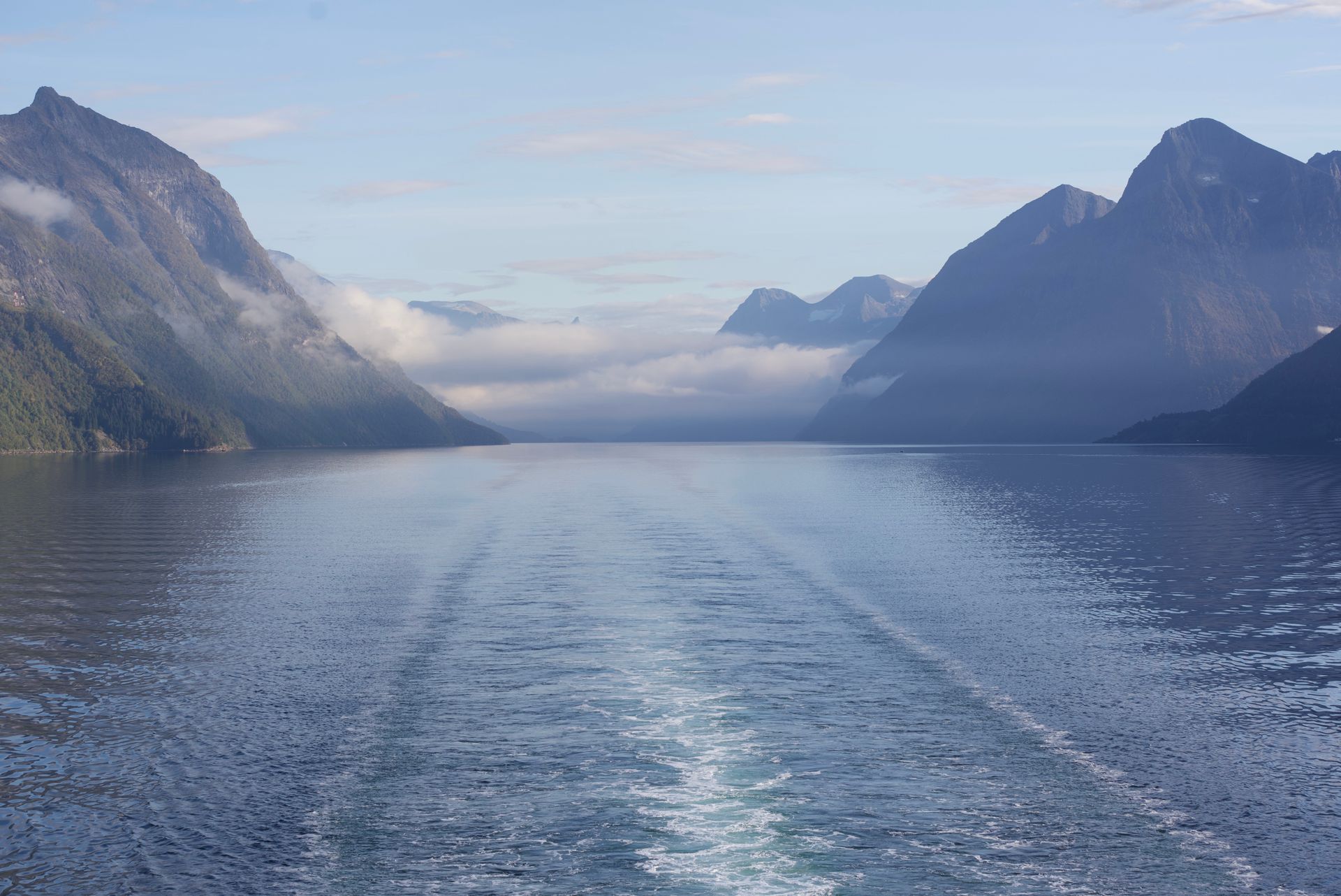 Calm, blue water with ship wake, flanked by steep, forested mountains, and low clouds, sunny sky.