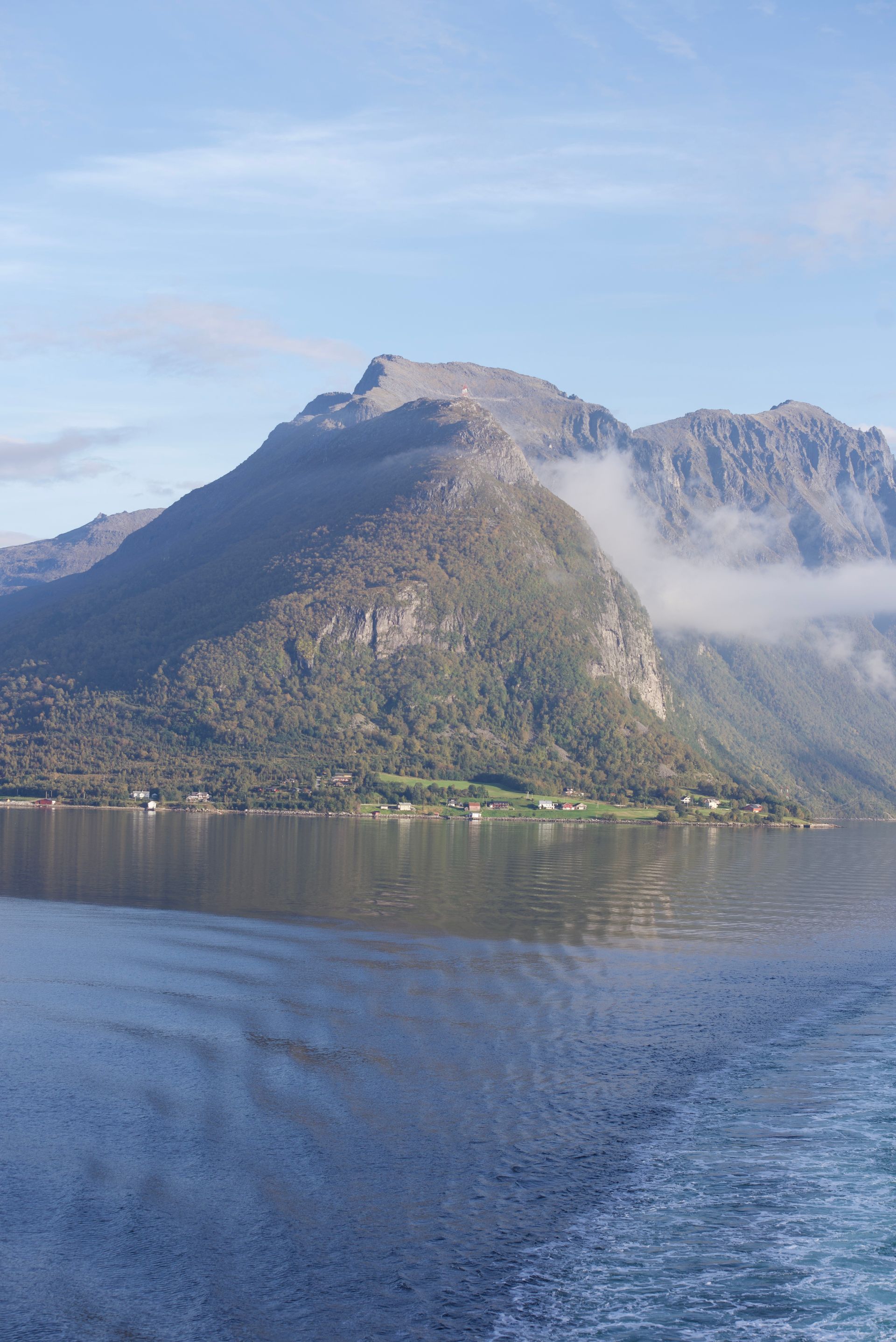 Mountains rising from a body of water, partially obscured by low-lying clouds; small buildings at the base.