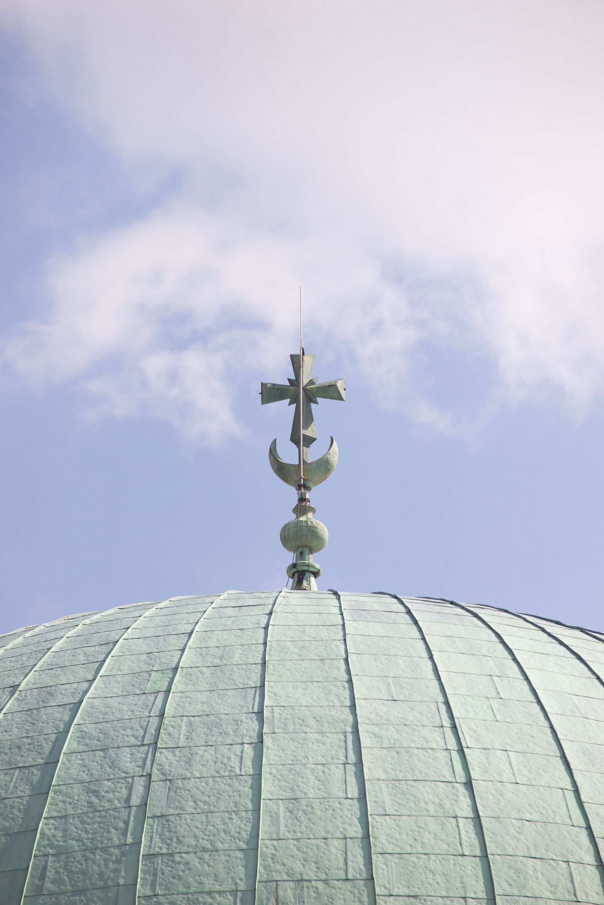 Green dome topped with a religious symbol combining a cross, crescent, and sphere, against a blue sky.